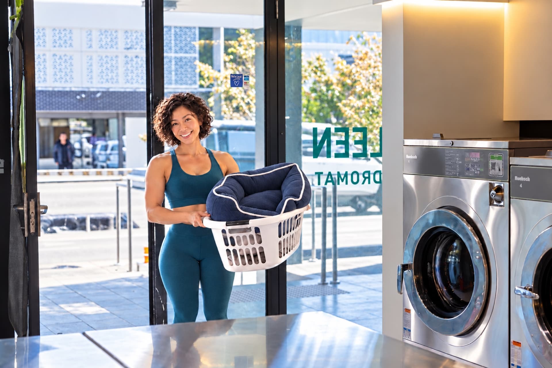 Customer carrying a laundry basket of pet bedding through the door of a Spakleen laundromat towards the folding bench and washing machines